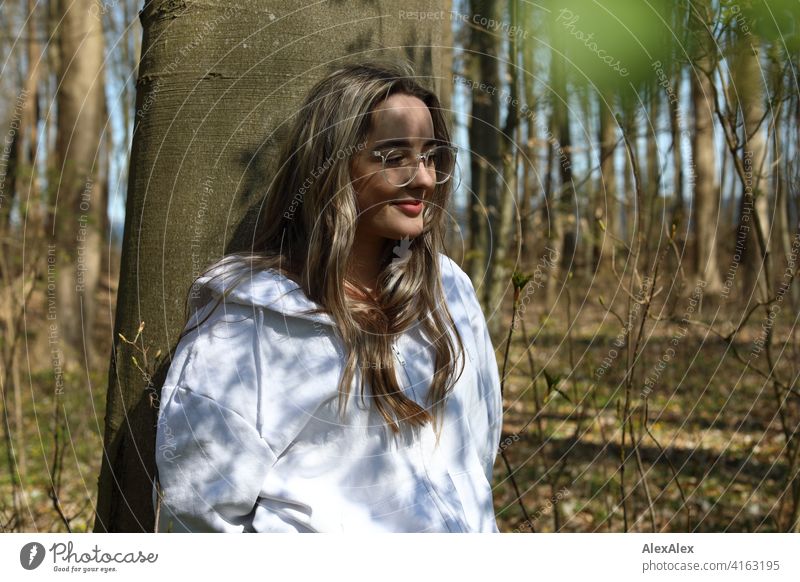 Ein schönes blondes Mädchen steht in einem Wald an einen Baum angelehnt und sieht seitlich in die Kamera, auf ihrem Gesicht ist ein Spiel aus Licht und Schatten von der Sonne und den Bäumen