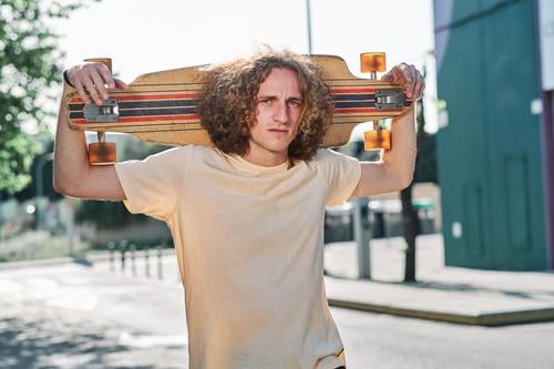 junger Mann mit lockigem Haar, der mit seinem Longboard oder Skeateboard auf den Schultern mitten auf der Straße in die Kamera schaut che Skateboard attraktiv