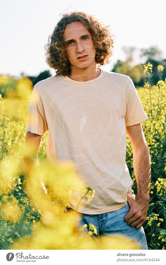 junger Mann mit lockigem Haar schaut in die Kamera in einem Rapsfeld an einem sonnigen Frühlingstag gelb Natur Landschaft che männlich Porträt Typ gutaussehend