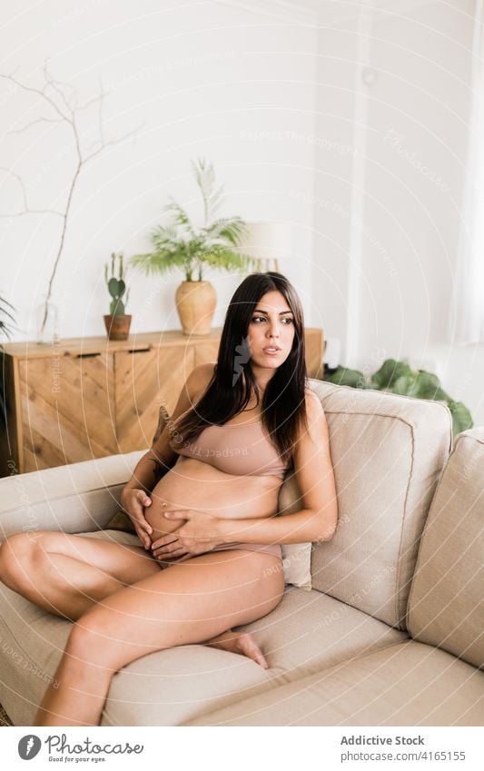 Lächelnde schwangere Frau auf dem Sofa zu Hause berühren Bauch Schwangerschaft gemütlich Dessous Unterwäsche sanft filigran Liege Mutter Pflege Angebot sitzen