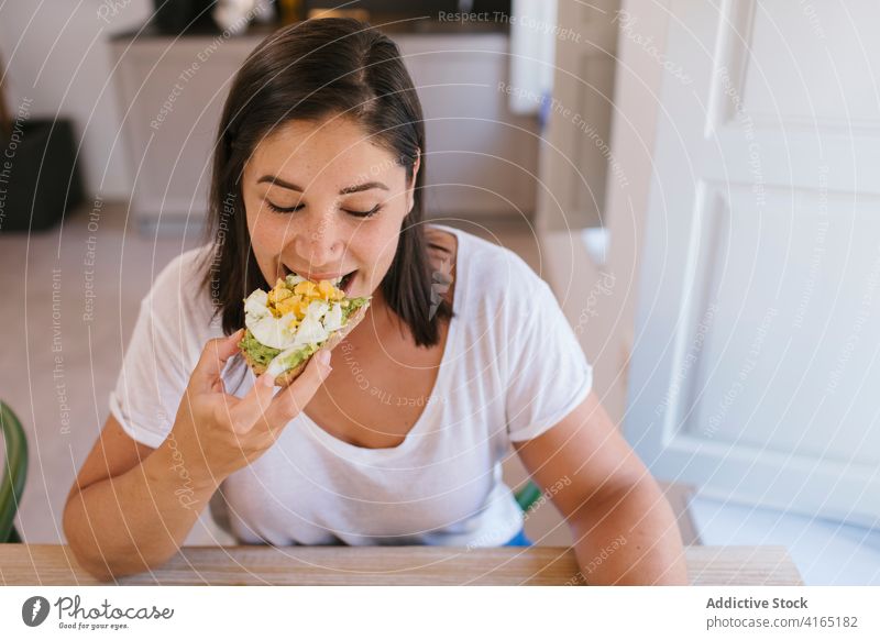 Frau beißt in einen Frühstückstoast im Innenbereich heimwärts Haus Mädchen Avocado kaufen Küche Kaffee Morgen Kaukasier brünett Tisch Lifestyle lässig Essen
