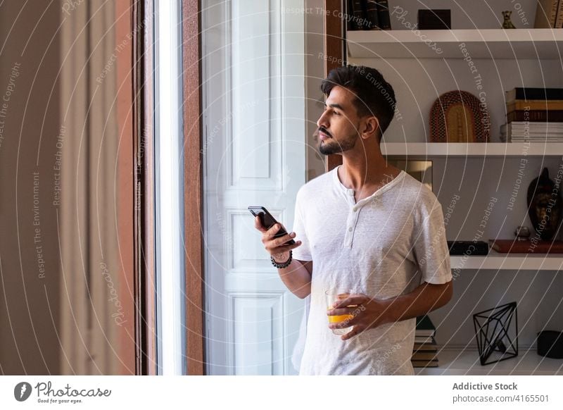 Junger Mann mit einem Glas Saft, der zu Hause auf seinem Smartphone surft Morgen Fenster benutzend trinken Browsen Apparatur Lifestyle jung männlich Vollbart