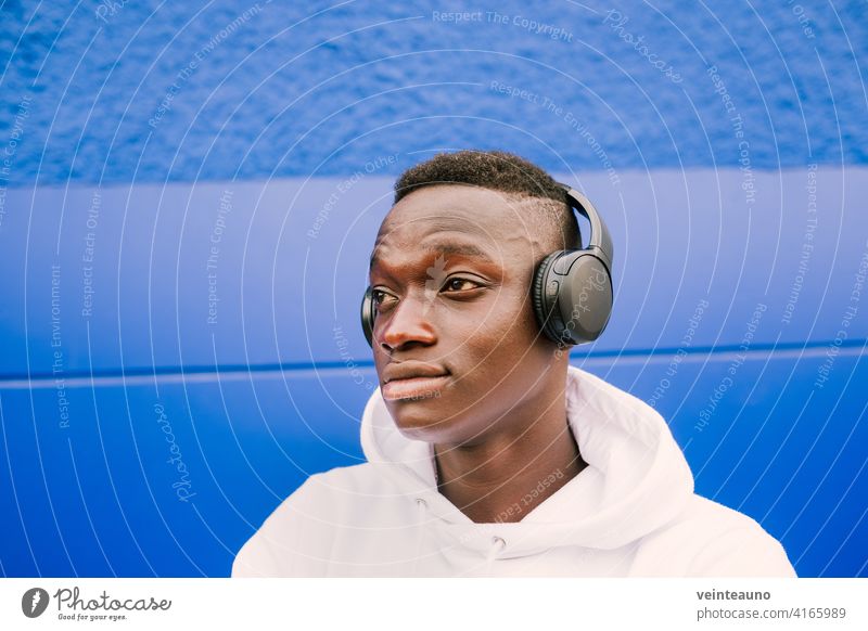 Junger afroamerikanischer schwarzer Mann, der Musik mit drahtlosen Kopfhörern hört, während er ein weißes Sweatshirt trägt und zur Seite schaut jung Afrikanisch