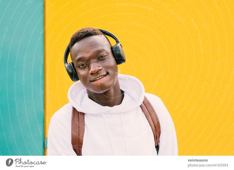 Junger afroamerikanischer schwarzer Mann, der mit drahtlosen Kopfhörern Musik hört, während er ein weißes Sweatshirt und einen Rucksack trägt und zur Kamera auf einem grünen und gelben Wandhintergrund schaut