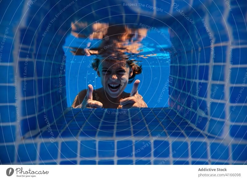 Aufgeregter Junge zeigt Daumen nach oben durch Poolwasser aufgeregt Daumen hoch gestikulieren Spaß haben Wasser sorgenfrei heiter unter Wasser Aktivität Lächeln