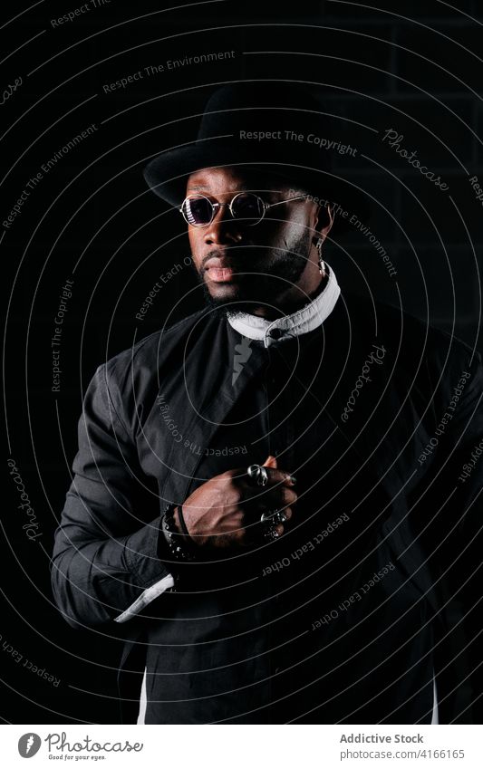 Ruhiger stilvoller Mann in dunklem Studio Stil schwarz Stoff Atelier Ring Hut Bekleidung trendy Sonnenbrille männlich ethnisch Afroamerikaner rund Form ernst