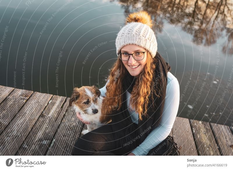 Junge Frau mit Brille sitzt mit einem kleinen Hund auf einem Holzsteg am See jung lachen sitzen Winter kalt Mütze Lebensfreude Freundschaft Zusammenhalt