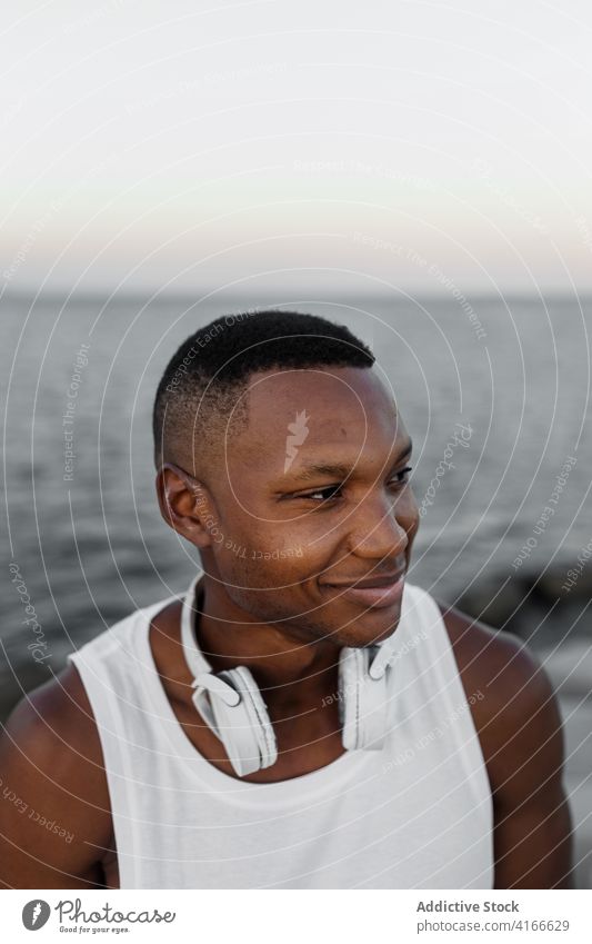 Fröhlicher ethnischer Mann lächelnd an der Küste des Meeres stehend aufgeregt Lächeln Kopfhörer positiv sorgenfrei Ufer genießen MEER Glück heiter männlich