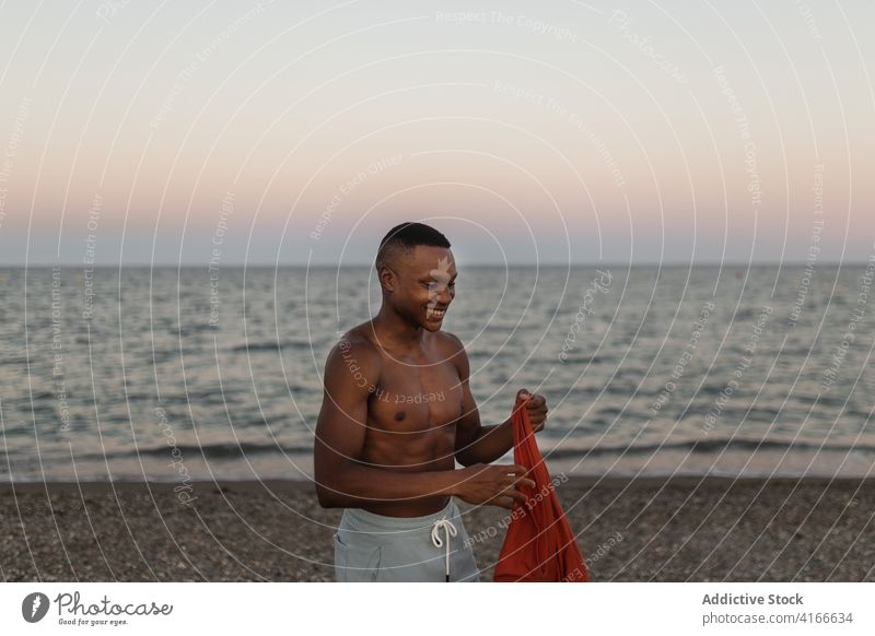 Lächelnder schwarzer Mann beim Anziehen eines T-Shirts am Strand Glück angezogen ruhen froh Athlet Küste sorgenfrei Meeresufer männlich Läufer MEER Lifestyle