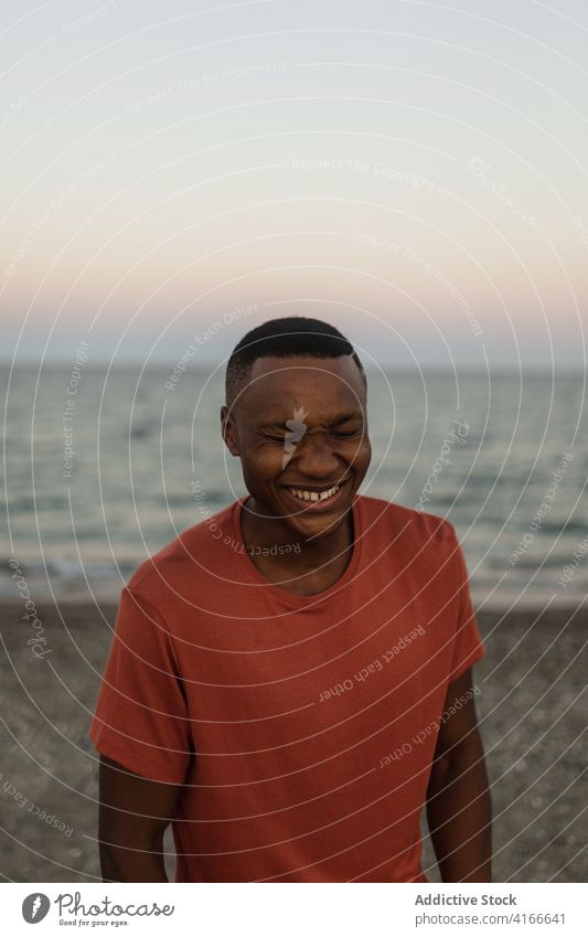 Fröhlicher schwarzer Sportler am Strand stehend Mann Afrikanisch Lächeln geschlossene Augen passen stark männlich sportlich ernst Typ Muskel Ufer selbstbewusst