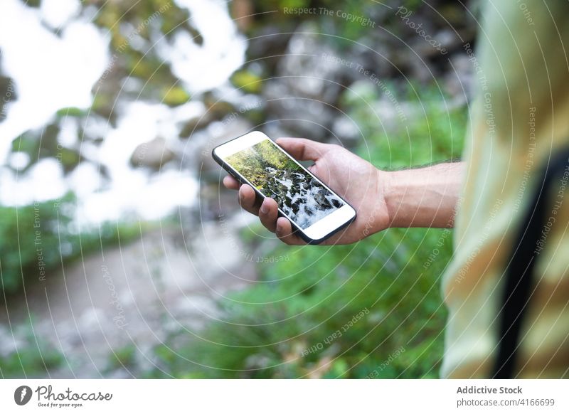 Anonyme männliche Person steht in der Nähe eines reißenden Flusses im Wald und macht ein Foto mit dem Mobiltelefon Reisender Mann Smartphone Urlaub Bach Tal rau