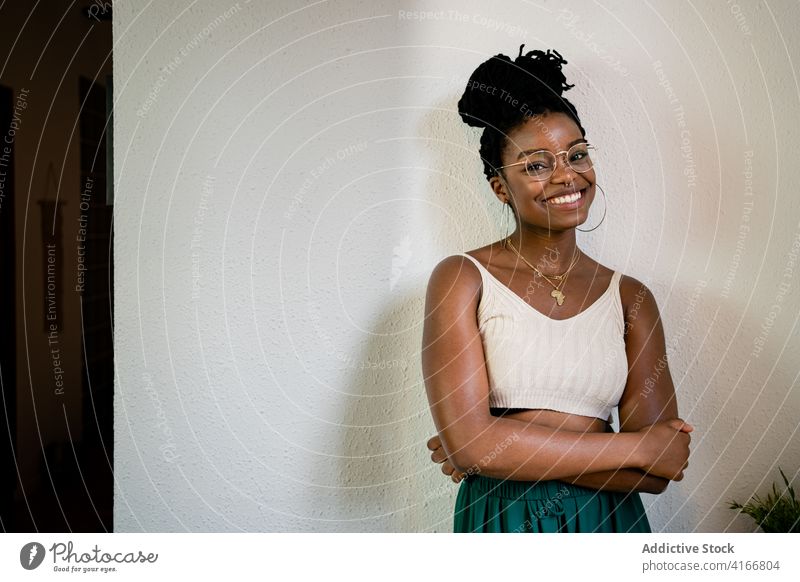 Glückliche schwarze Frau mit Brille im Zimmer Zahnfarbenes Lächeln Frisur Accessoire Mode Outfit Stil Inhalt heiter Raum Afroamerikaner brünett Freude positiv