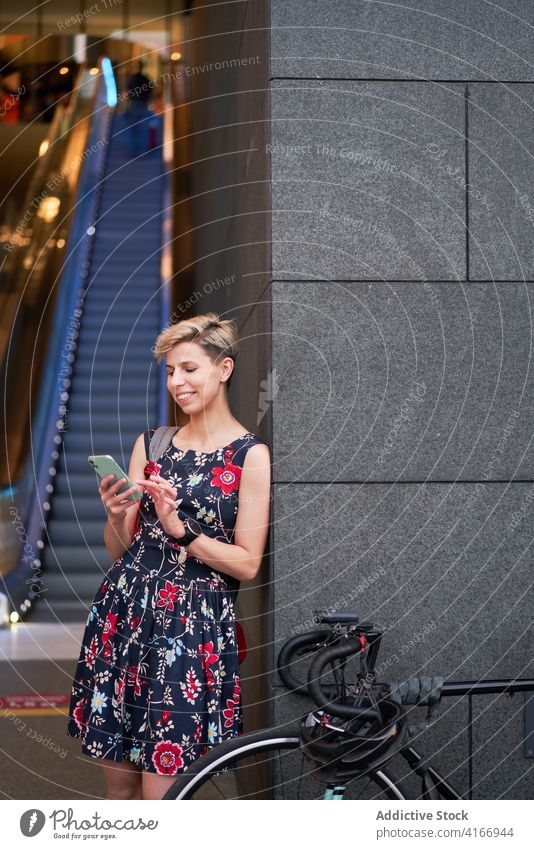 Junge Frau mit Telefon in den Händen steht in der Nähe von modernen Gebäude, Rolltreppe Kleid Fahrrad Abend Spaziergang Mädchen jung Reiten schön Großstadt