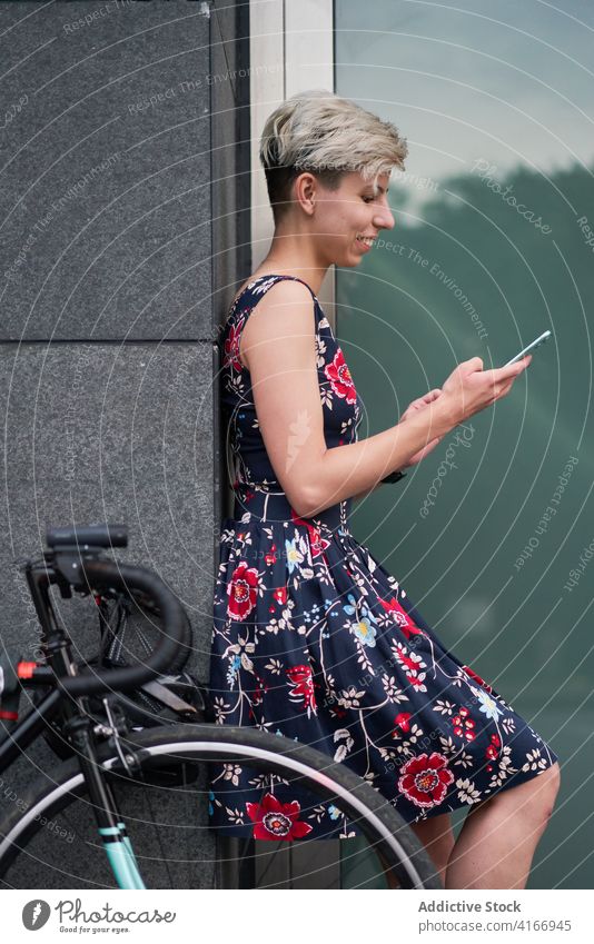 Seitenansicht einer Frau mit Telefon in den Händen steht in der Nähe eines modernen Gebäudes Kleid Fahrrad Abend Spaziergang Mädchen jung Reiten schön Großstadt