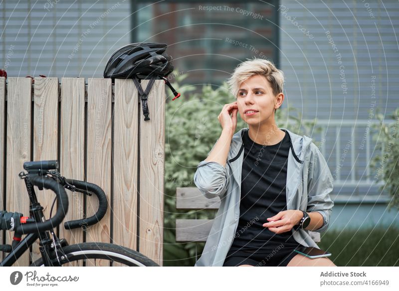 Weibliche Kurierin telefoniert, während sie auf einer Bank neben einem Fahrrad im Hintergrund eines Gebäudes sitzt Radfahrer Großstadt Tag Frau jung Sport