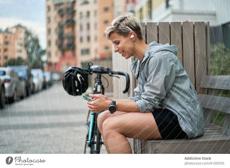 Seitenansicht einer glücklichen Kurierin mit Telefon in den Händen, die auf einer Bank neben einem Fahrrad vor einem Gebäude sitzt Radfahrer Großstadt Tag Frau