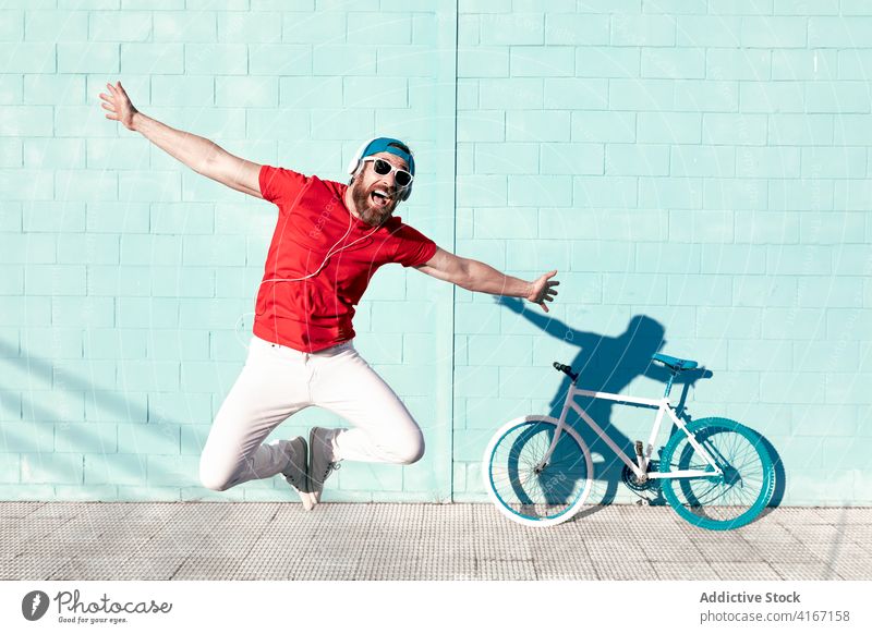 Fröhlicher junger Mann springt auf der Straße in der Nähe von modernen Fahrrad und blauen Gebäude springen ausdehnen heiter expressiv schreien trendy urban