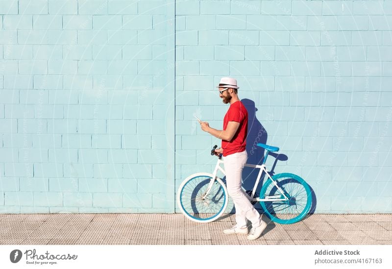 Trendy junger Mann mit Smartphone in der Nähe von Fahrrad auf der Straße benutzend Gebäude Großstadt Streetstyle trendy Fahrzeug Sonnenlicht Mobile Telefon