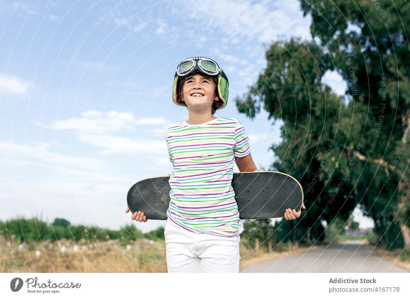 Glücklicher verträumter Junge mit Skateboard auf der Fahrbahn Kind Schutzbrille Landschaft stylisch Bekleidung wolkig Blauer Himmel Kindheit Straße positiv