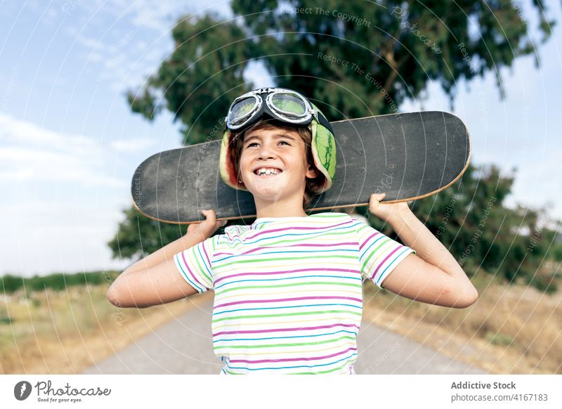 Glücklicher verträumter Junge mit Skateboard auf der Fahrbahn Kind Schutzbrille Landschaft stylisch Bekleidung wolkig Blauer Himmel Kindheit Straße positiv