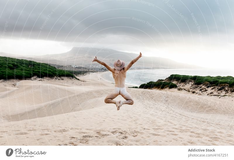 Anonymer sportlicher nackter Mann, der an der sandigen, einsamen Meeresküste springt jum aktiv sorgenfrei Freiheit Strand malerisch reisen Meeresufer Hut passen