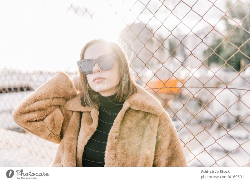 Stilvolle Frau in warmer Jacke in der Stadt Fell trendy Outfit Bestimmen Sie Sonnenbrille Oberbekleidung Netz Zaun Straße urban cool Mode ernst Windstille