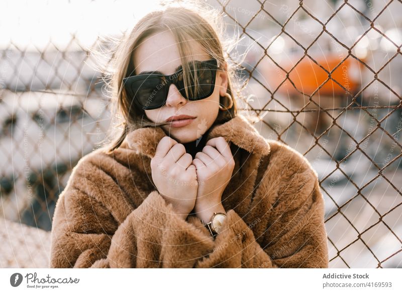 Stilvolle Frau in warmer Jacke in der Stadt Fell trendy Outfit Bestimmen Sie Sonnenbrille Oberbekleidung Netz Zaun Straße urban cool Mode ernst Windstille