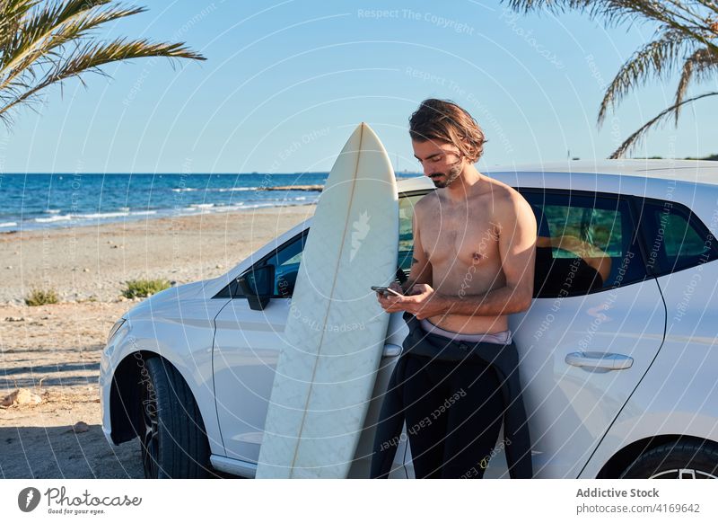Mann ohne Hemd mit Surfbrett in der Nähe des Autos und Smartphone Surfer Browsen Strand PKW benutzend soziale Netzwerke Sommer männlich passen Holzplatte Surfen