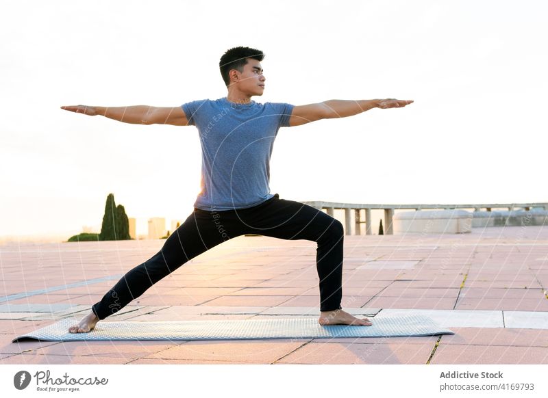 Ethnischer Mann in Sportkleidung macht Yoga bei Sonnenuntergang Krieger-Pose üben Asana beweglich Fokus ruhig männlich ethnisch asiatisch virabhadrasana urban