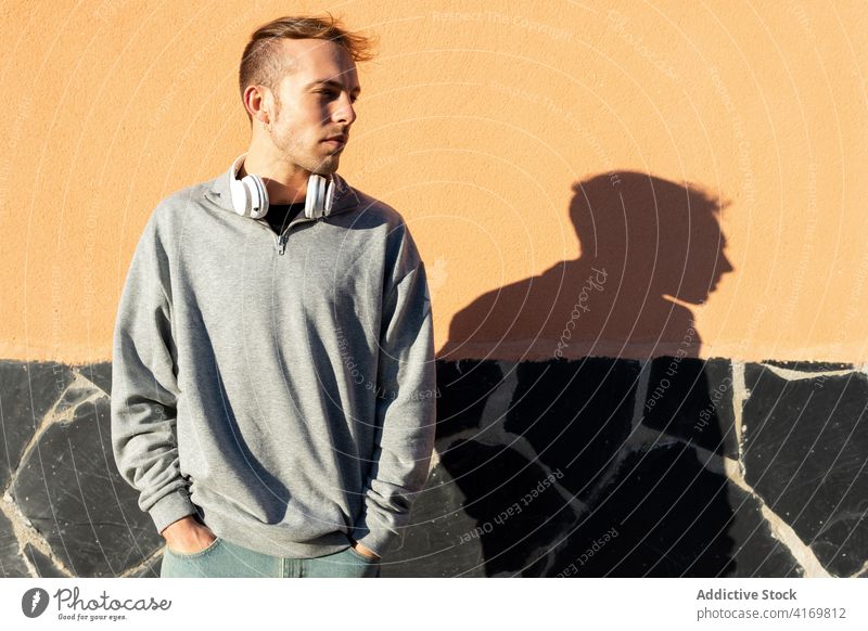 Junger Mann mit Kopfhörer schaut weg Sonnenlicht Sonnenschein Deckung behüten ausdehnen Hipster urban männlich modern unrasiert lässig Wand Straße Lifestyle
