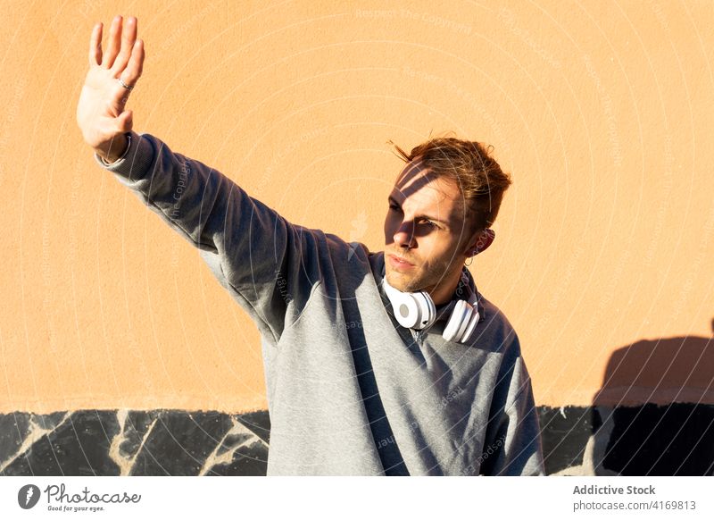 Junger Mann mit Kopfhörern, die das Gesicht vor Sonnenlicht schützen Sonnenschein Deckung behüten ausdehnen Hipster urban männlich modern unrasiert lässig Wand