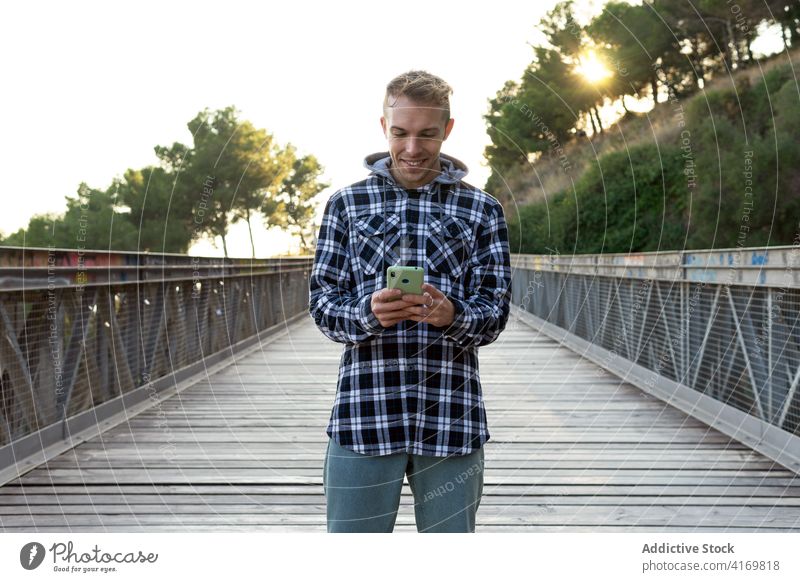 Glücklicher Mann mit Mobiltelefon auf einem Steg stehend Smartphone benutzend Brücke Hipster positiv prüfen Reisender Telefon jung männlich Landschaft Apparatur