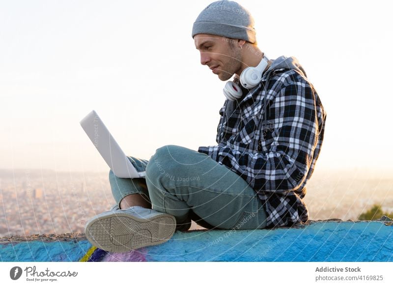 Positiver junger Mann in Freizeitkleidung mit Laptop auf der Straße benutzend Hipster Glück positiv Browsen online Internet Blogger freiberuflich