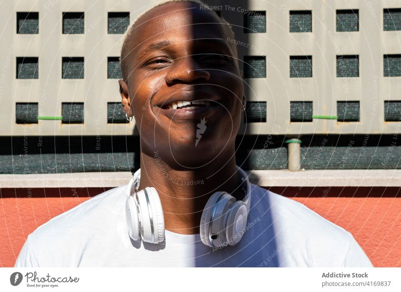 Lächelnder ethnischer Mann mit Kopfhörern in der Stadt urban cool Straße Großstadt genießen sonnig Wochenende Schatten männlich schwarz Afroamerikaner Gebäude
