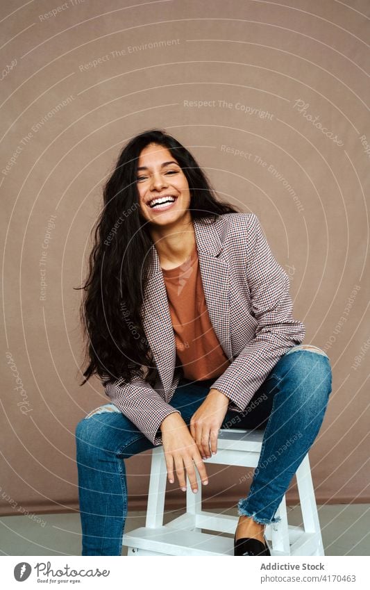 Glückliche ethnische Frau sitzt auf einem Stuhl im Studio trendy smart lässig Stil Model Jacke Lächeln Outfit heiter Atelier selbstbewusst Lachen sitzen