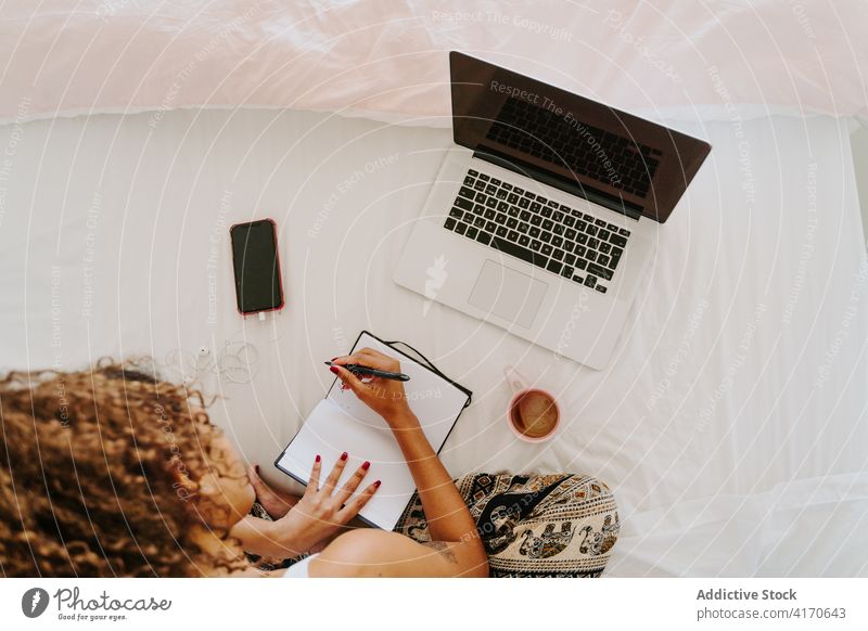 Nicht erkennbare ethnische Frau mit Laptop und Planer bei der Arbeit im Schlafzimmer zu Hause Bett zur Kenntnis nehmen freiberuflich abgelegen lässig jung