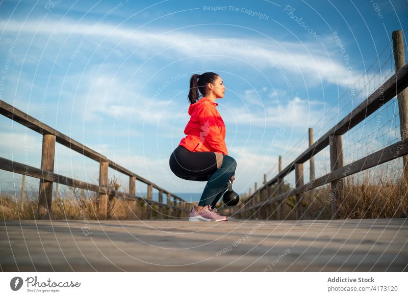 Sportlerin macht Übungen mit einer Kettlebell auf einem Holzsteg Kniebeuge Training Athlet Ausdauer Spazierweg Natur Frau Sportbekleidung physisch hölzern