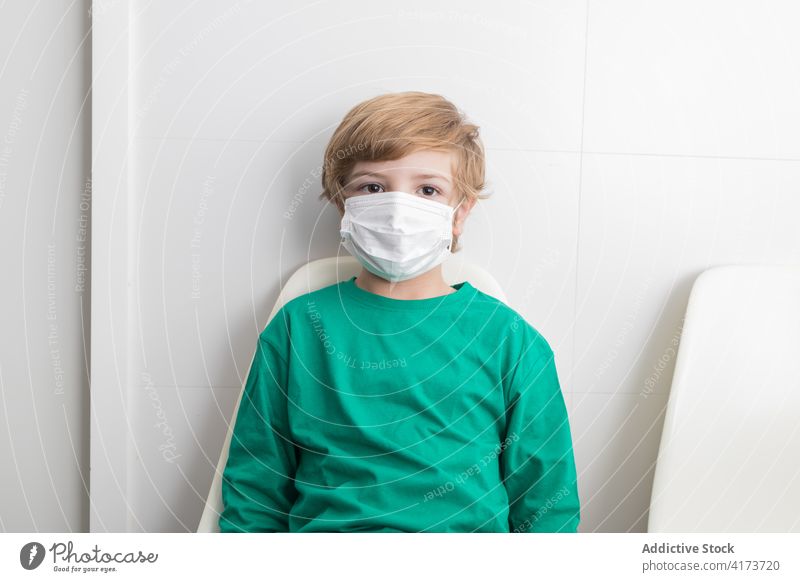 Kind mit medizinischer Maske im Krankenhaus Mundschutz Coronavirus Bund 19 Junge behüten Klinik Stuhl Raum sitzen Windstille COVID19 Seuche verhindern Ausbruch