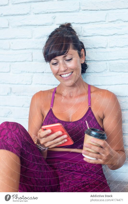 Fröhliche Frau benutzt Smartphone während einer Yogastunde Pause benutzend fettarm Backsteinwand Lächeln trinken zum Mitnehmen Erwachsener Lektion Browsen Gerät