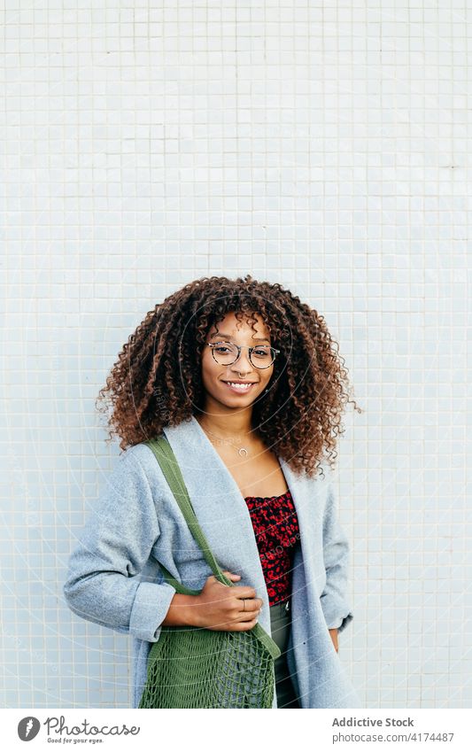 Afroamerikanische Frau in der Nähe einer gefliesten Wand fettarm Straße ruhen Stil Lächeln Großstadt Outfit lässig Tasche Baumwolle trendy Glück krause Haare