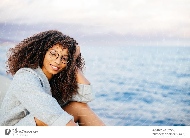 Fröhliche schwarze Frau sitzt auf Böschung Stauanlage MEER Lächeln Stil Wochenende Glück Outfit ruhen jung Kai heiter ethnisch Afroamerikaner sitzen Wasser