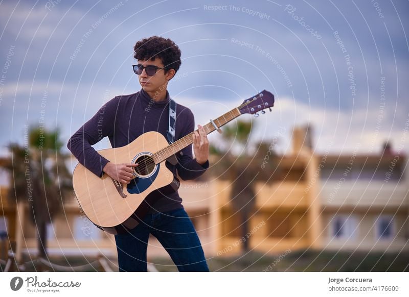 junge kaukasische Teenager-Junge spielt akustische Gitarre am Strand. Sonnenbrille und dunkle Kleidung. Kunst Künstler Blues lässig Kaukasier Komponist Konzert