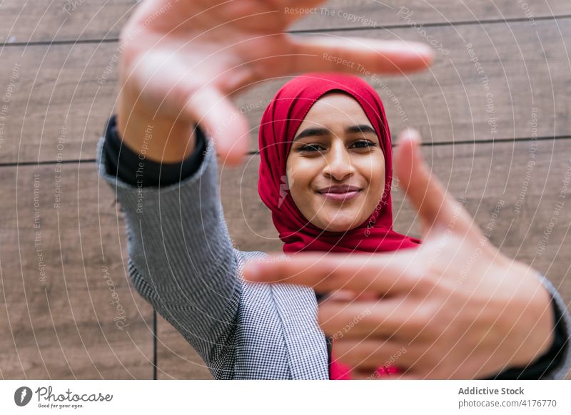 Lächelnde ethnische Frau mit Kopftuch zeigt rahmende Geste Rahmen gestikulieren einfangen Zeichen Hijab muslimisch Foto arabisch Optimist Glück heiter Ernte