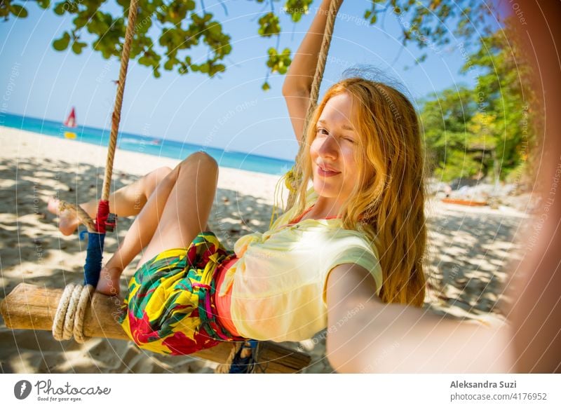 Glückliche junge Frau mit roten Haaren lachen und schwingen auf Schaukel auf einem Baum am Strand, ein Selfie zu nehmen. Schöne Sommer sonnigen Tag, türkisfarbenes Meer, weißer Sand, tropische Landschaft. Phuket, Thailand.