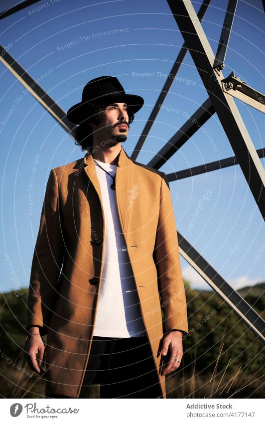 Stilvoller Mann in Hut und Mantel steht in der Nähe von Metallturm trendy selbstbewusst Turm modern ernst jung Vollbart männlich ethnisch Lifestyle Outfit