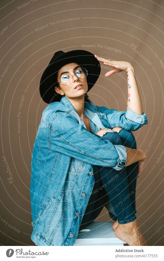 Stilvolle Frau in Jeans-Outfit und Hut im Studio Knie umarmend berühren trendy Jeansstoff Atelier Model Hocker sitzen Windstille Kleidungsstück Bekleidung