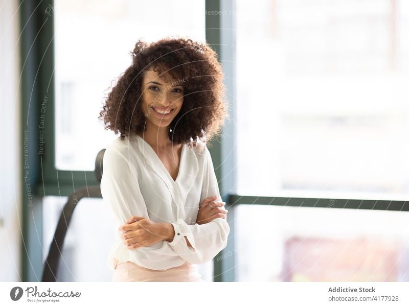 Lächelnde ethnische Frau, die in der Nähe eines Fensters im Büro steht heiter Glück jung positiv Erfolg Optimist hispanisch krause Haare modern Lifestyle froh