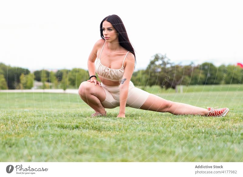 Schlanke Frau in modischer Sportkleidung, die ihre Beine im Gras ausstreckt Fitness Training Dehnung passen sportlich trendy Stil Park Sportbekleidung Übung