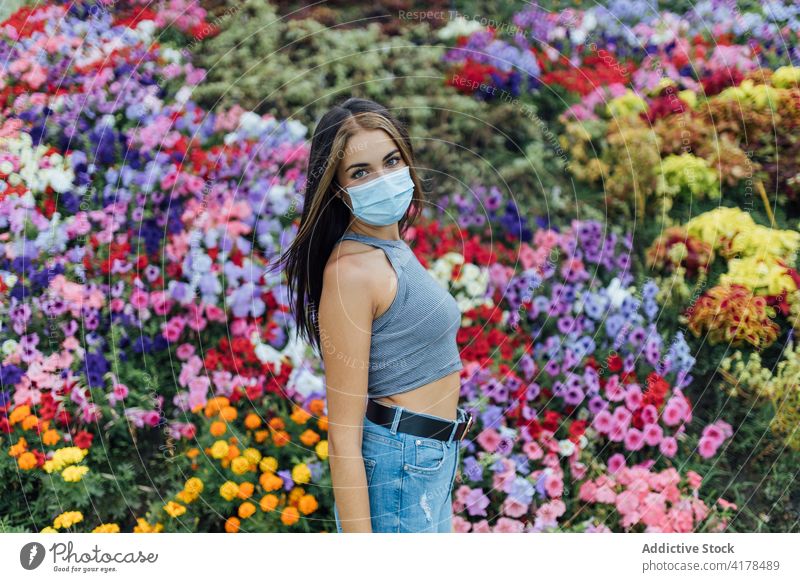 Junge Frau in medizinischen Maske stehen in der Nähe von blühenden Blumen Mundschutz behüten Park Coronavirus Bund 19 Pandemie neue Normale jung lässig