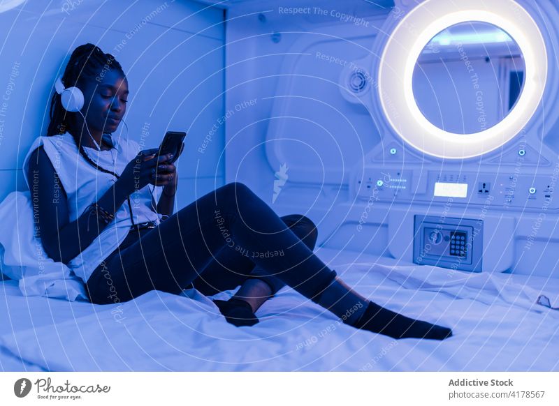 Ethnische Frau benutzt Smartphone in einem Kapselhotel Hotel benutzend ruhen Reisender Bett Kopfhörer Browsen Mobile Telefon zuhören Apparatur Gerät online